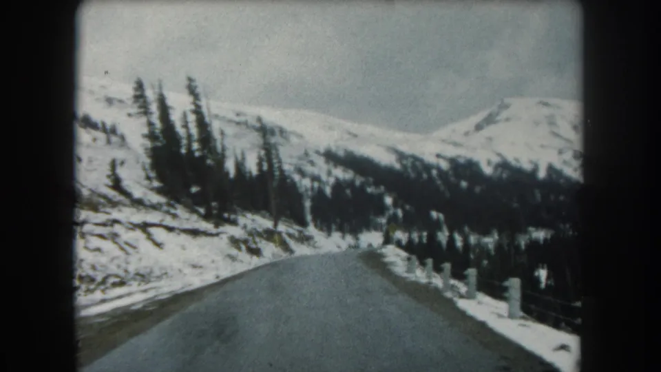 1962 8mm view of a paved Summit County mountain road bordered by snowbanks, dark evergreens, and distant snowy ridges.