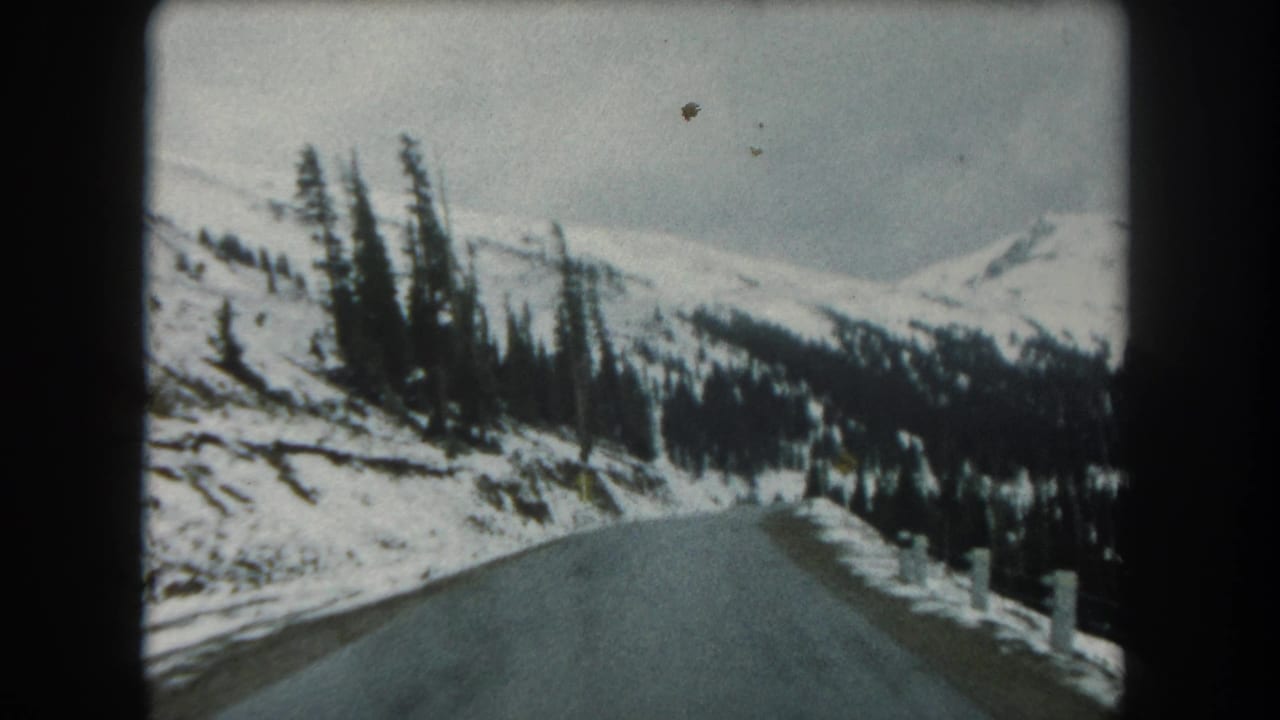 Summit County winter drive frame showing a paved road climbing through snow-covered forested terrain under a gray sky.