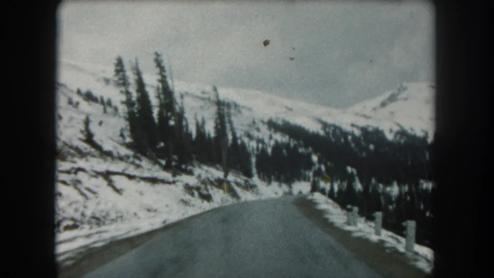 Summit County winter drive frame showing a paved road climbing through snow-covered forested terrain under a gray sky.