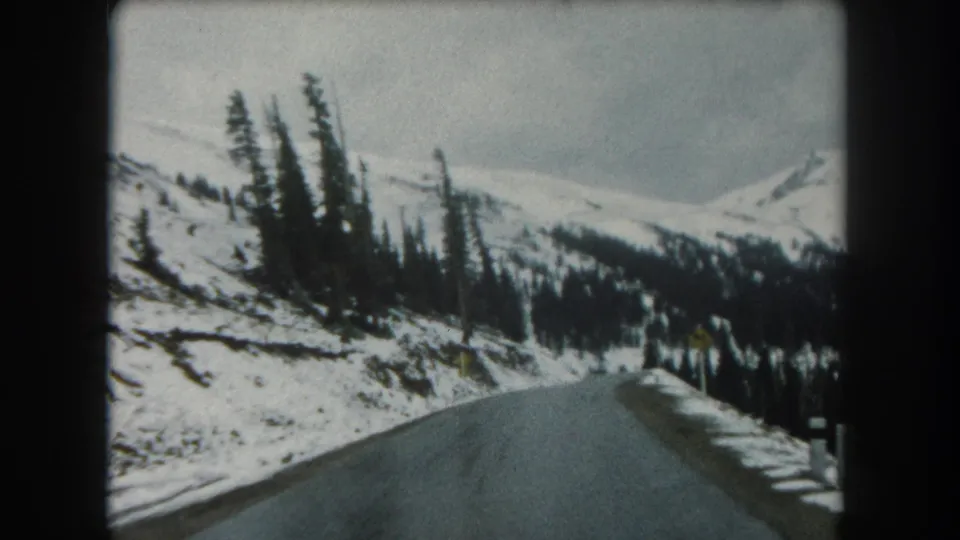 1960s home-movie frame of a Colorado mountain road bending through snow-covered roadside and evergreen forest.