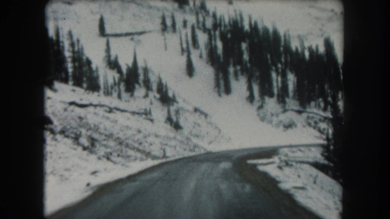 Moving 8mm travel view of a wet one-lane road beside snow and conifers in Summit County, Colorado, in 1962.