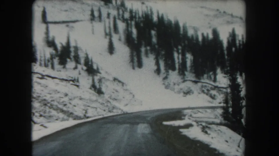 Soft 8mm frame showing a wet mountain road beneath snow-covered slopes and dense evergreen trees in 1962 Colorado footage.