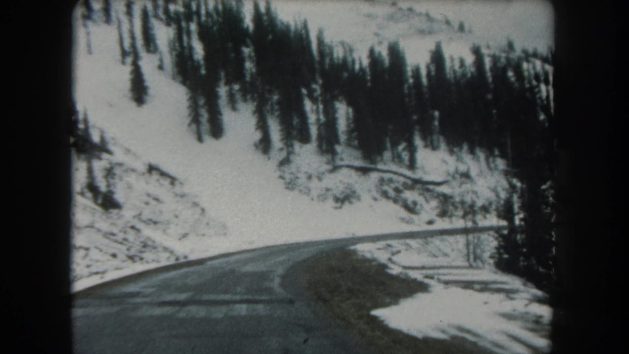 1962 8mm view of a curving Summit County, Colorado mountain road bordered by snow, trees, and rocky hillside.