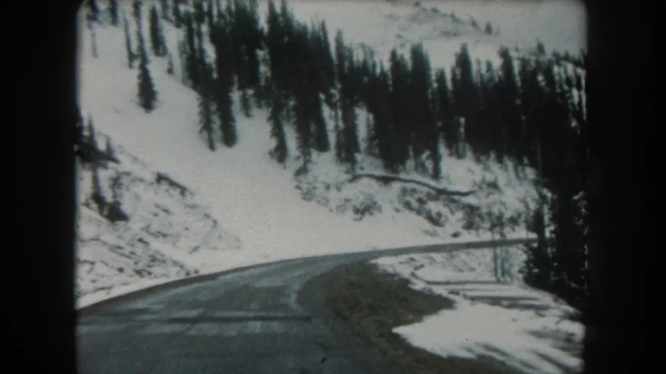 1962 8mm view of a curving Summit County, Colorado mountain road bordered by snow, trees, and rocky hillside.