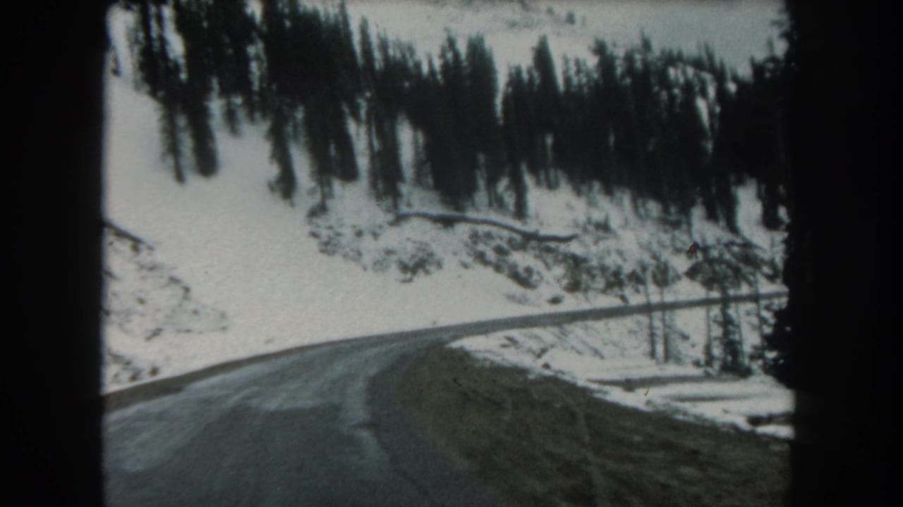 Forward-facing 1962 home-movie frame of a snowy mountain road in Summit County, Colorado, with trees and hillside above.