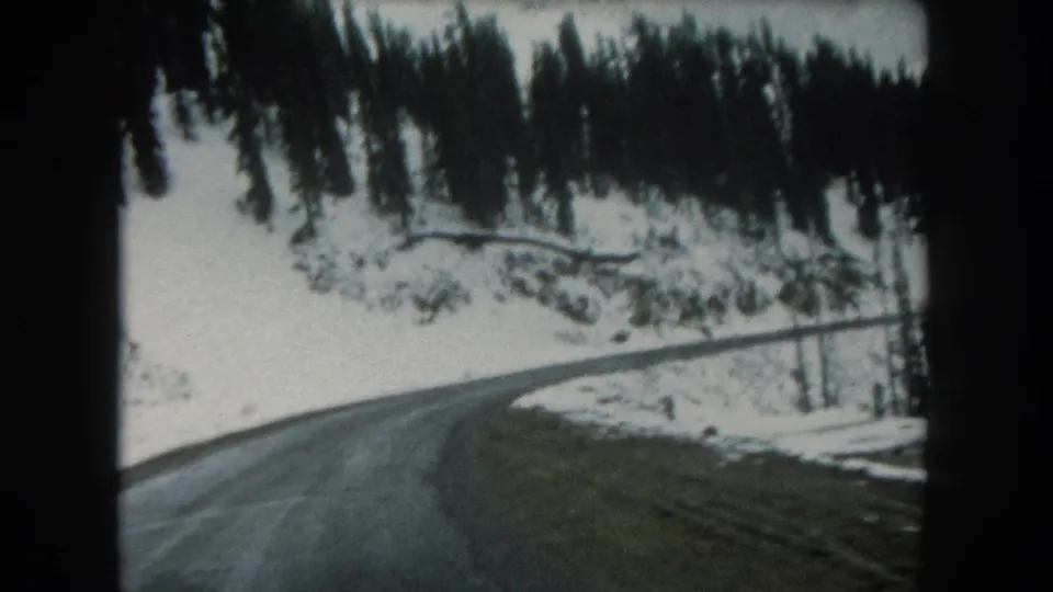 1962 8mm passenger-view frame of a mountain road bend in Summit County, Colorado, with snowbanks and evergreen trees.
