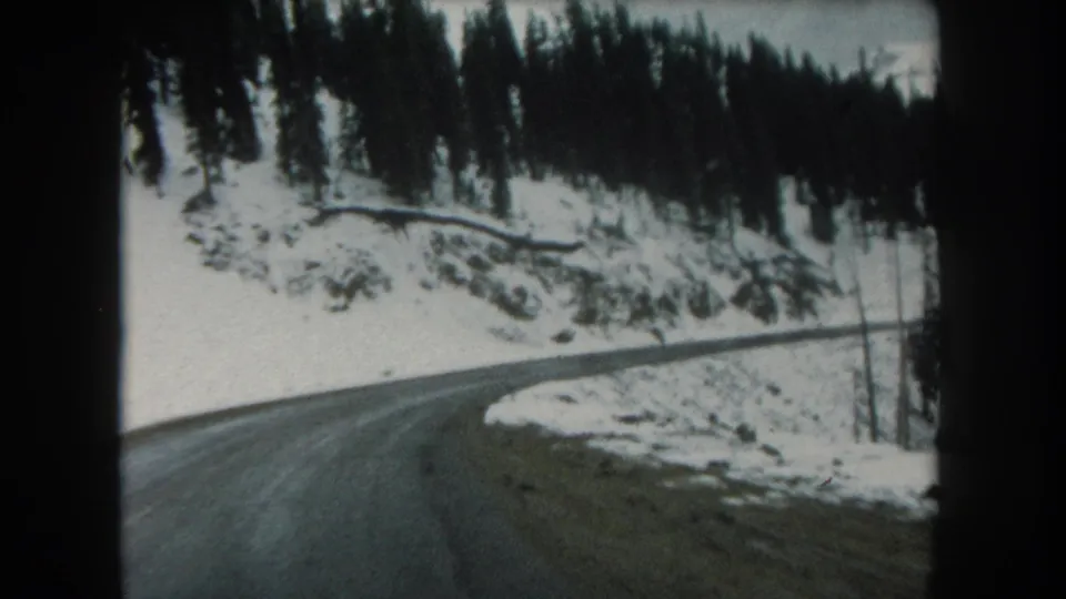Late frame from a 1962 Summit County, Colorado road sequence showing asphalt, snow, evergreens, and a mountain slope.