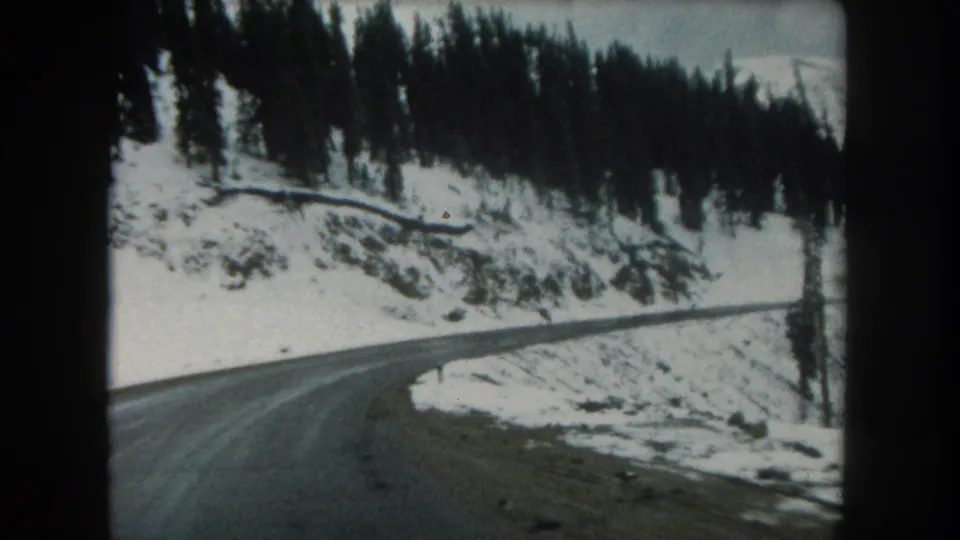 1962 Summit County, Colorado 8mm frame showing a wet road bending through snowy evergreen-covered hills.