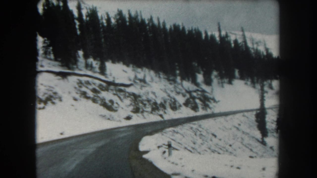1962 Summit County travel frame with a wet mountain road, snow-covered shoulder, and dense evergreens along the slope.