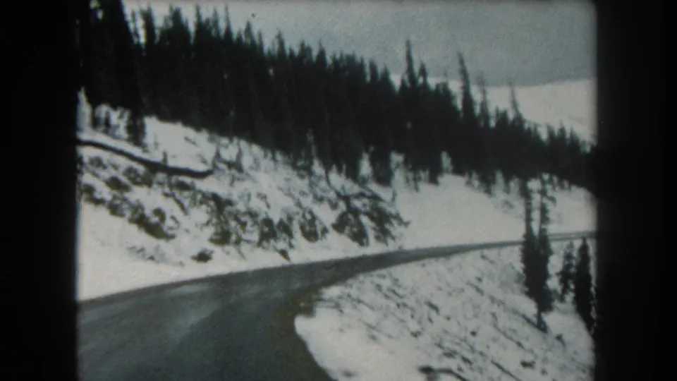 Passenger-view 8mm frame from Summit County, Colorado, looking along wet pavement beside snow and dark conifers.
