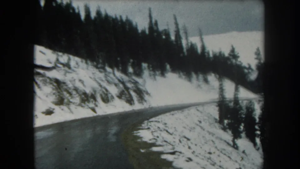1962 Colorado mountain-road frame showing wet asphalt curving through snowbanks and evergreen trees under gray light.
