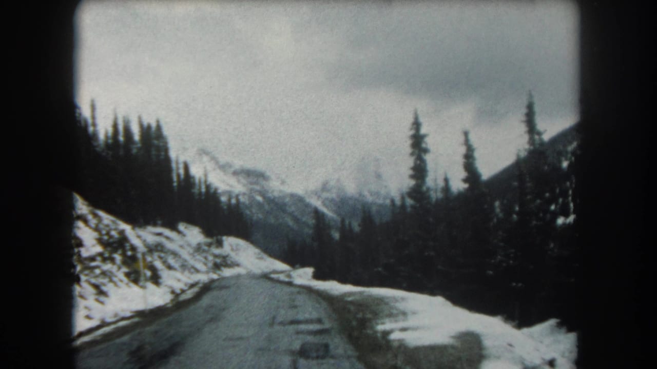 1962 Summit County, Colorado road view with snowbanks, evergreen trees, wet pavement, and distant mountain slopes under an overcast sky.