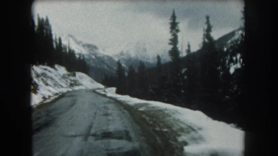 1962 Summit County, Colorado driving view looking down a wet road between snow-covered shoulders and evergreen trees in the mountains.
