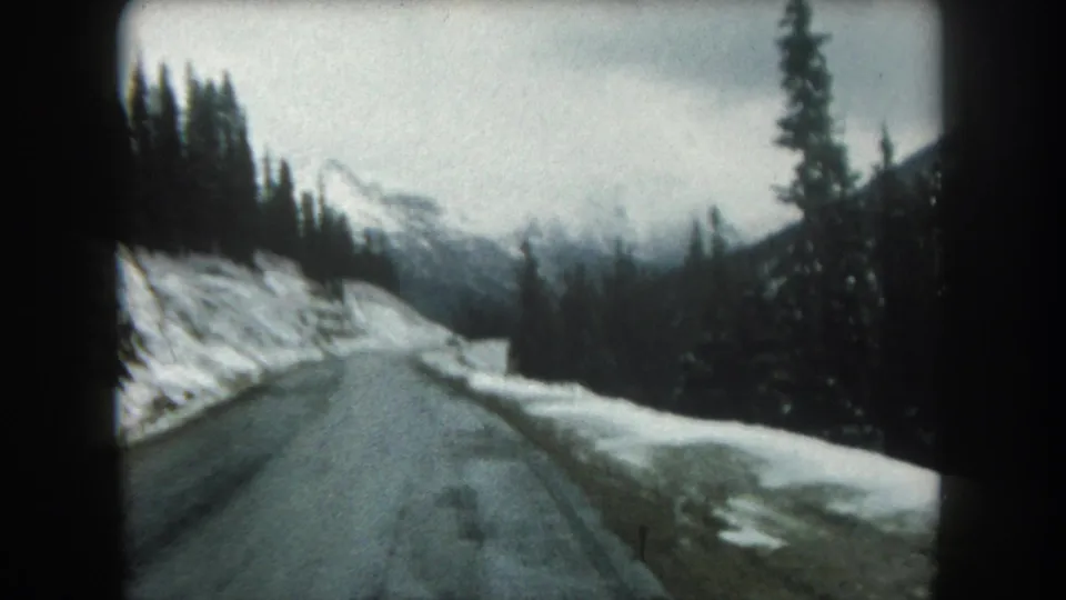 Final 1962 Summit County road frame showing a wet snowy roadway, pine trees, distant mountain forms, and soft 8mm color under cloudy light.