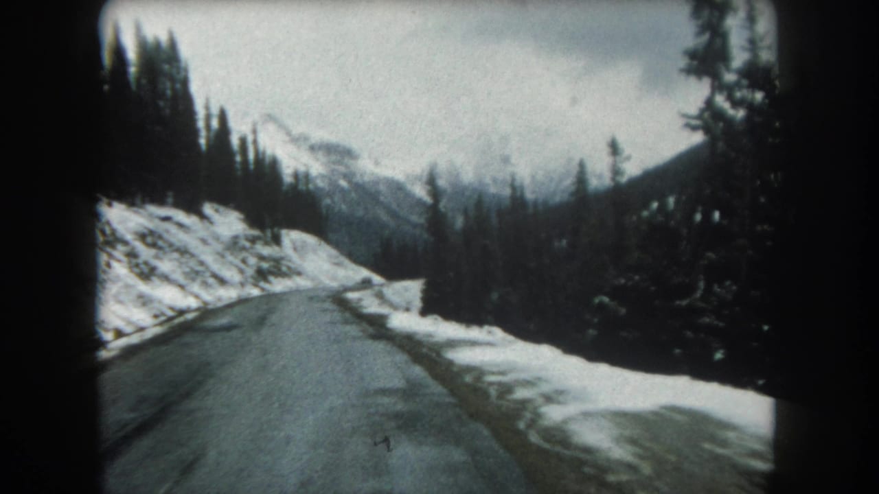 Snow-lined mountain road curving through evergreen forest with distant peaks under a cloudy sky.