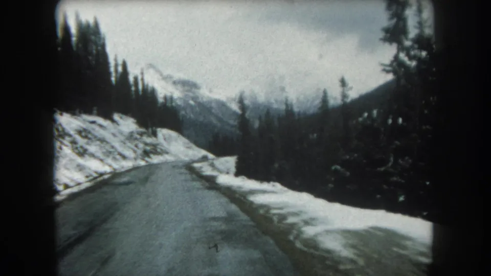 1962 Summit County, Colorado home-movie frame of a snowy mountain road bordered by evergreens and distant peaks.
