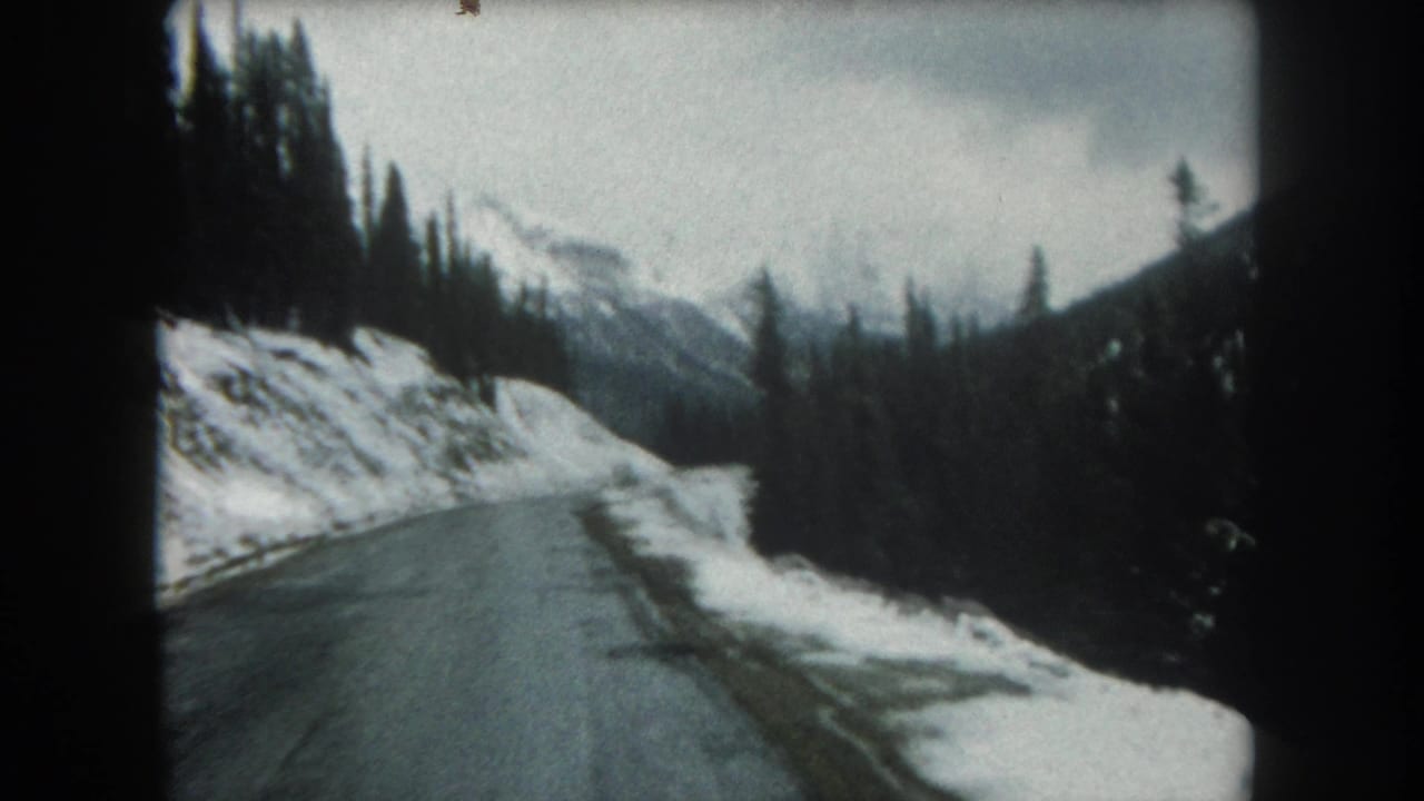 Mountain road bordered by snowbanks and dark evergreen trees, with peaks visible in the distance.