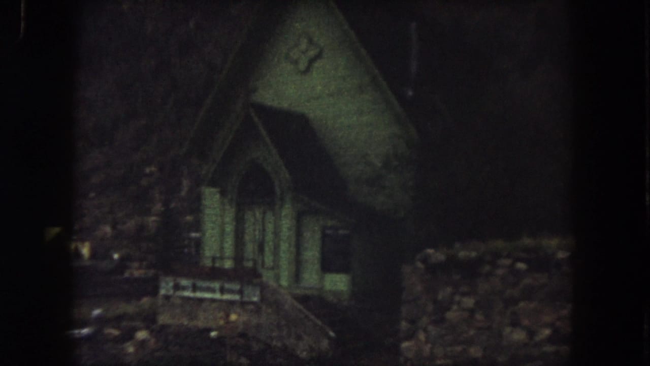 1962 Summit County 8mm frame showing a small dim house or cabin with a steep roof, arched doorway, and dark wooded surroundings.