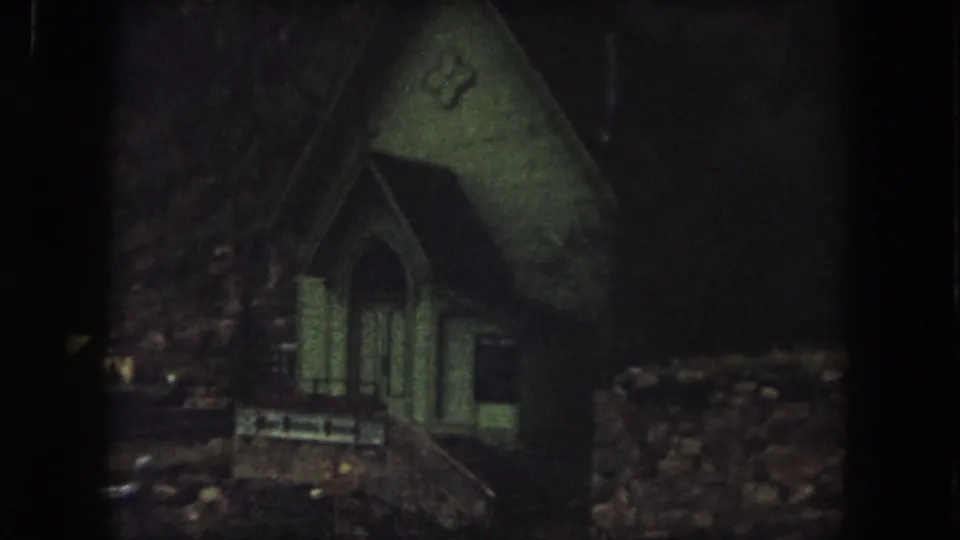 1962 Summit County 8mm frame showing a small dim house or cabin with a steep roof, arched doorway, and dark wooded surroundings.