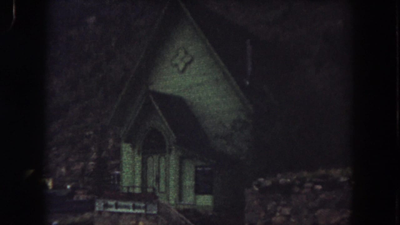 1962 Summit County view of a small green church with an arched entrance, cross emblem, and hillside surroundings.