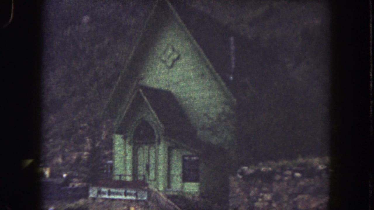 Green church with arched windows and cross emblem beside a low rough hillside.