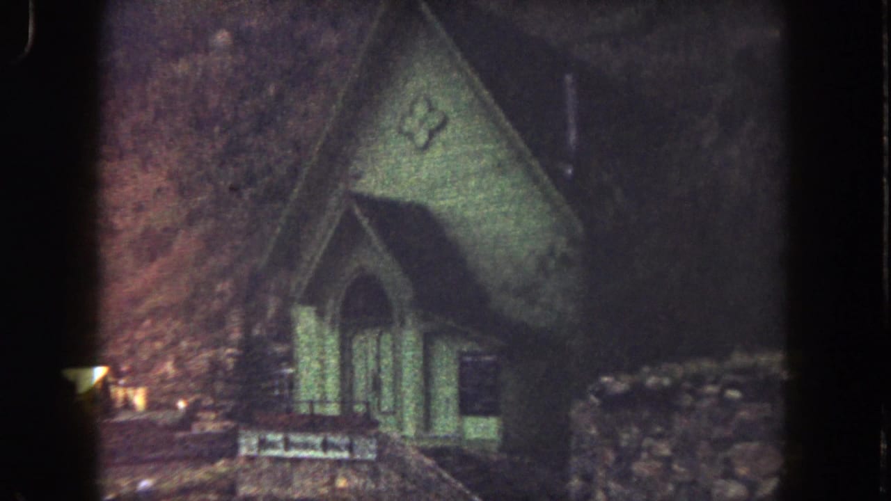 1962 home-movie frame showing the green church exterior, arched entrance, cross emblem, and hilly terrain.