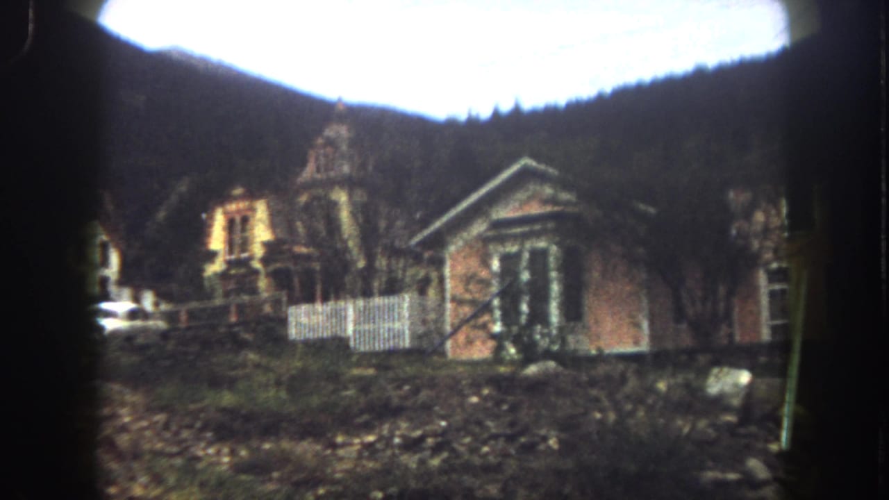 Wider 1962 Summit County view of rustic buildings, a white fence, rough ground, and forested mountain backdrop.