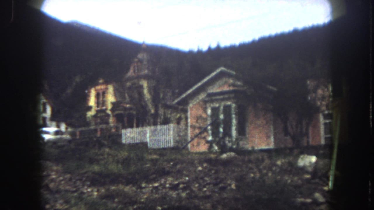 1962 8mm view of houses on a slope in Summit County, Colorado, with a white fence, rocky ground, trees, and wooded mountains behind.