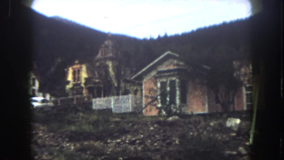 1962 8mm view of houses on a slope in Summit County, Colorado, with a white fence, rocky ground, trees, and wooded mountains behind.