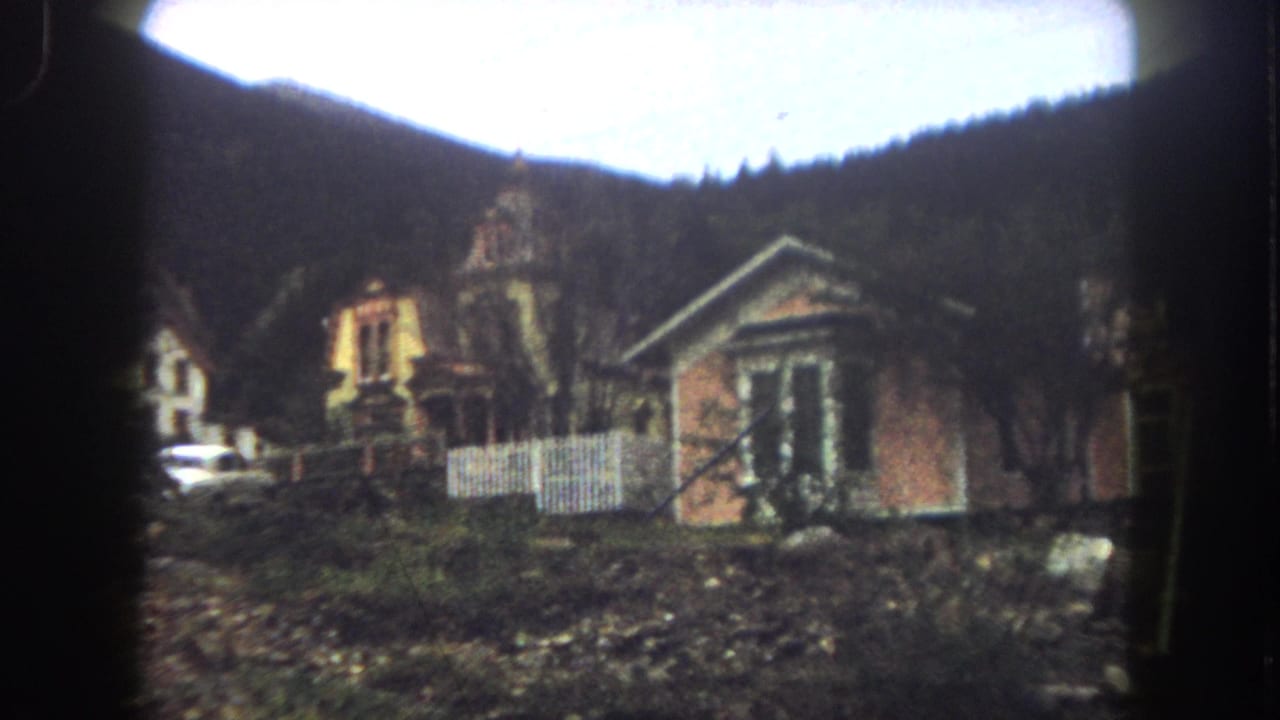Weathered hillside houses with a fence, bare trees, rocky ground, and wooded mountains behind.