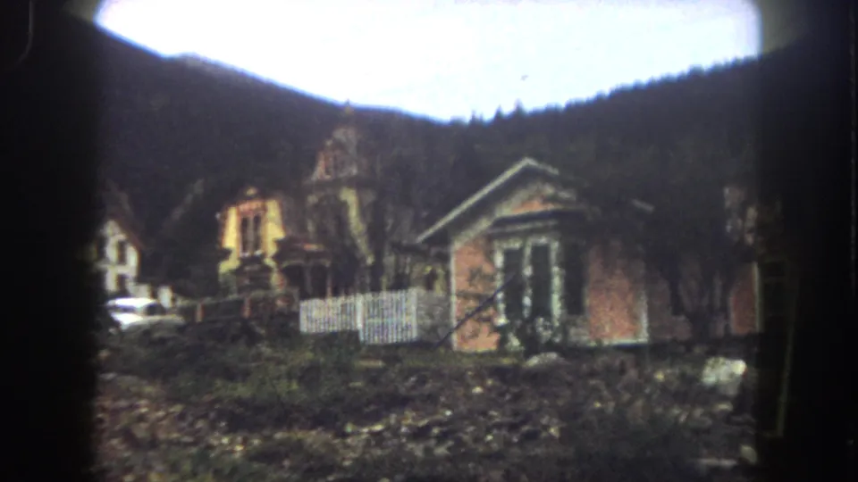 1962 Summit County, Colorado home-movie frame showing hillside houses, a white fence, a light car, bare trees, and dark mountain woods.