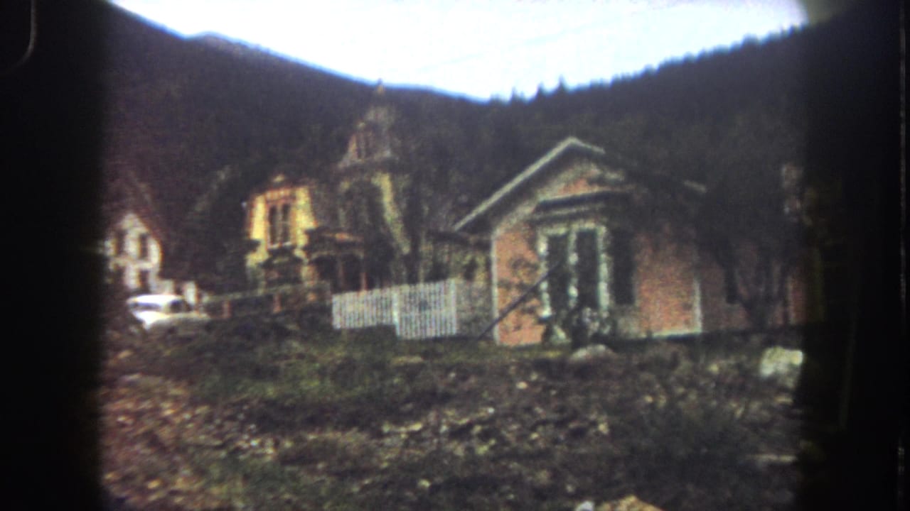 Summit County, Colorado 8mm frame from 1962 showing old hillside houses, a white picket fence, parked car, rough ground, and forested mountains.