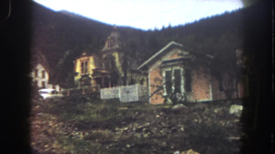 Summit County, Colorado 8mm frame from 1962 showing old hillside houses, a white picket fence, parked car, rough ground, and forested mountains.