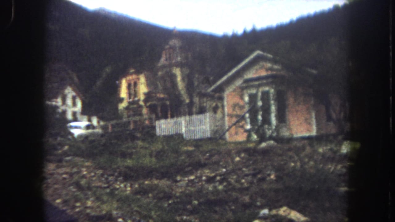 Final 1962 8mm frame of hillside houses in Summit County, Colorado, with a white fence, rocky foreground, parked car, and wooded mountain slope.
