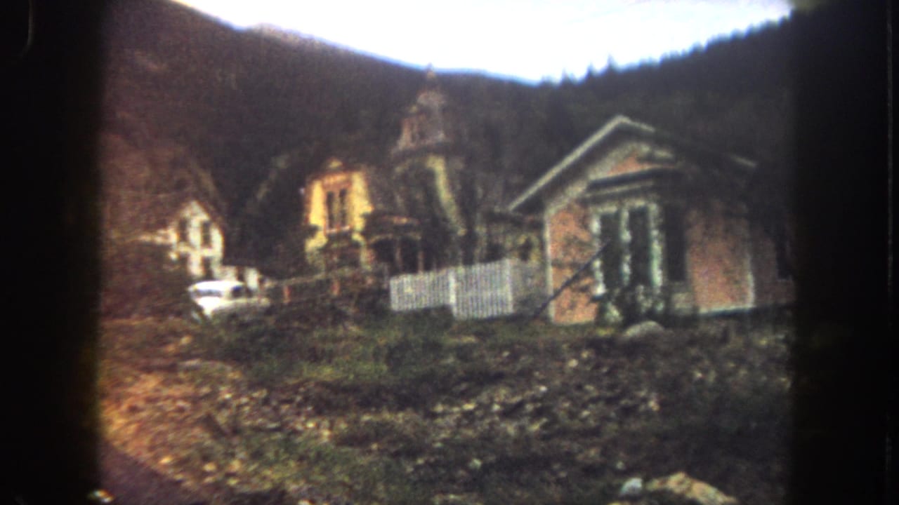 Small Victorian-style house exterior on a hillside with a white fence and parked car.