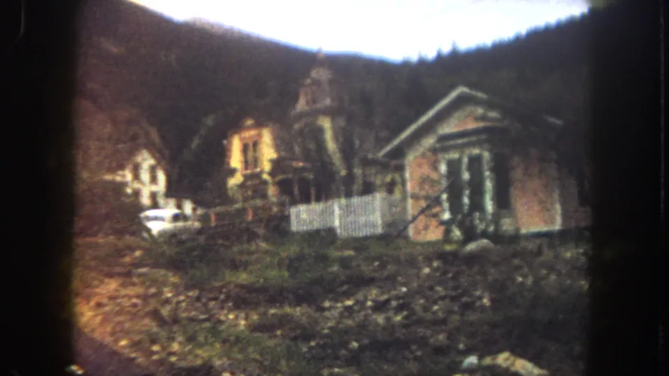 1962 8mm frame from Summit County, Colorado showing a Victorian-style house exterior near a hillside, with a white fence, stone wall, and parked white car partly visible.