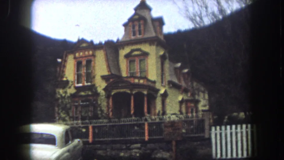 1962 8mm frame from Summit County, Colorado showing the front of a yellow Victorian-style house with turret, porch columns, fenced yard, and wooded hillside background.