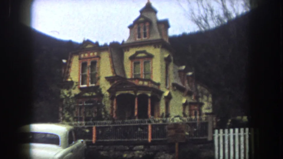 Final 1962 8mm frame of the Summit County, Colorado house exterior, showing the turreted yellow facade, porch, fence, white gate, parked car, and dark wooded slope behind.
