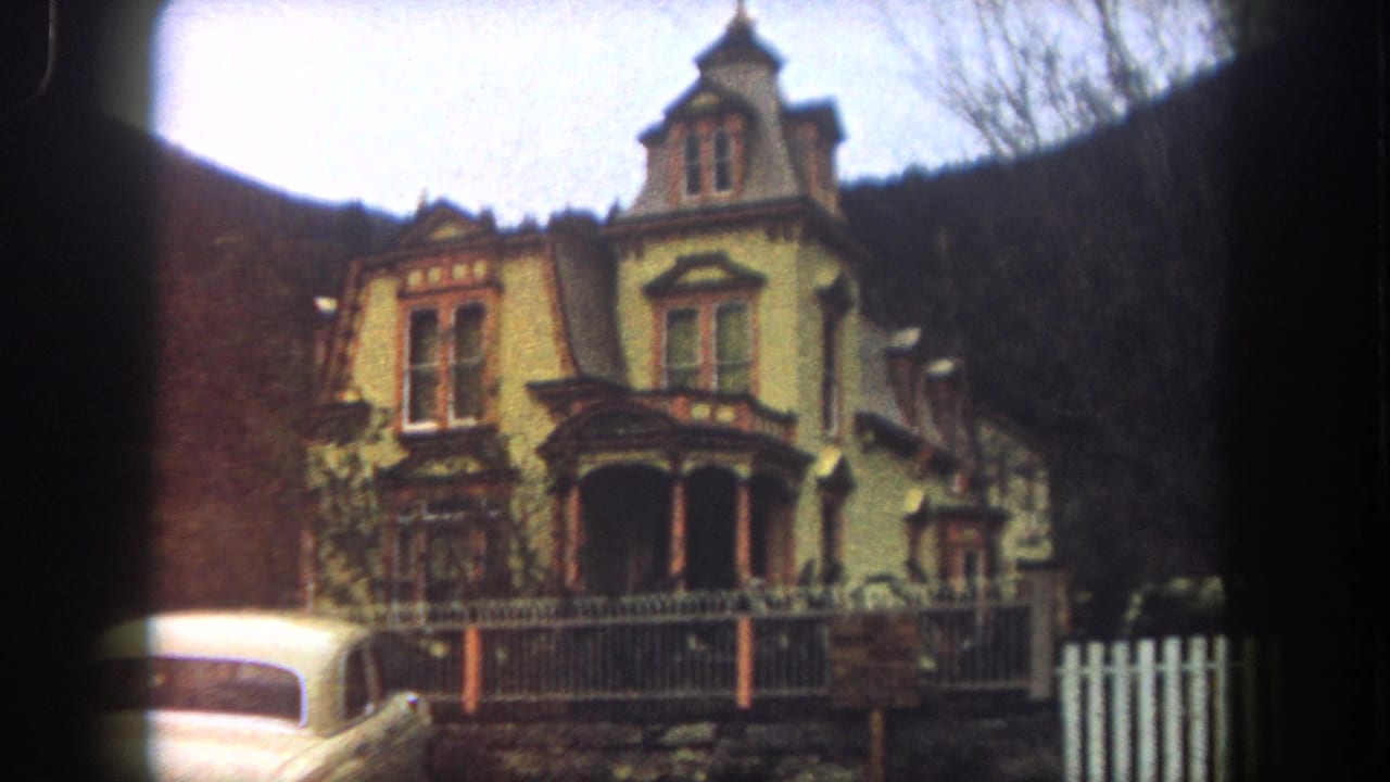 1962 8mm frame from Summit County, Colorado showing a yellow Victorian-style hillside house, porch, fence, gate, and white car.
