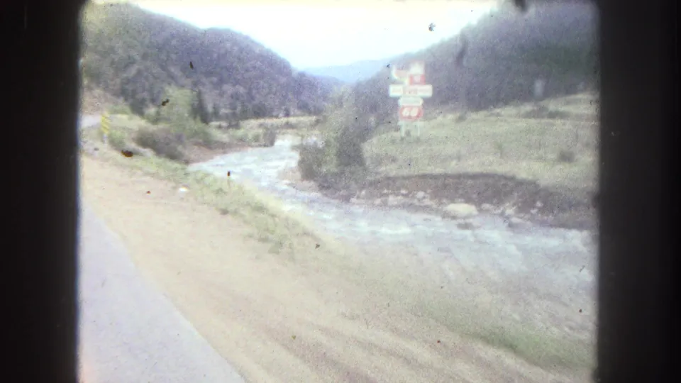 1962 8mm travel frame in Summit County, Colorado showing a mountain road, rushing stream, valley slopes, and a sign marked 66.