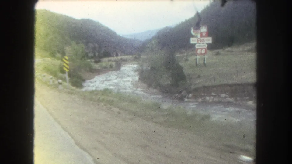 Summit County, Colorado home-movie frame from 1962 showing a stream, roadside shoulder, mountains, trees, and a 66 sign.