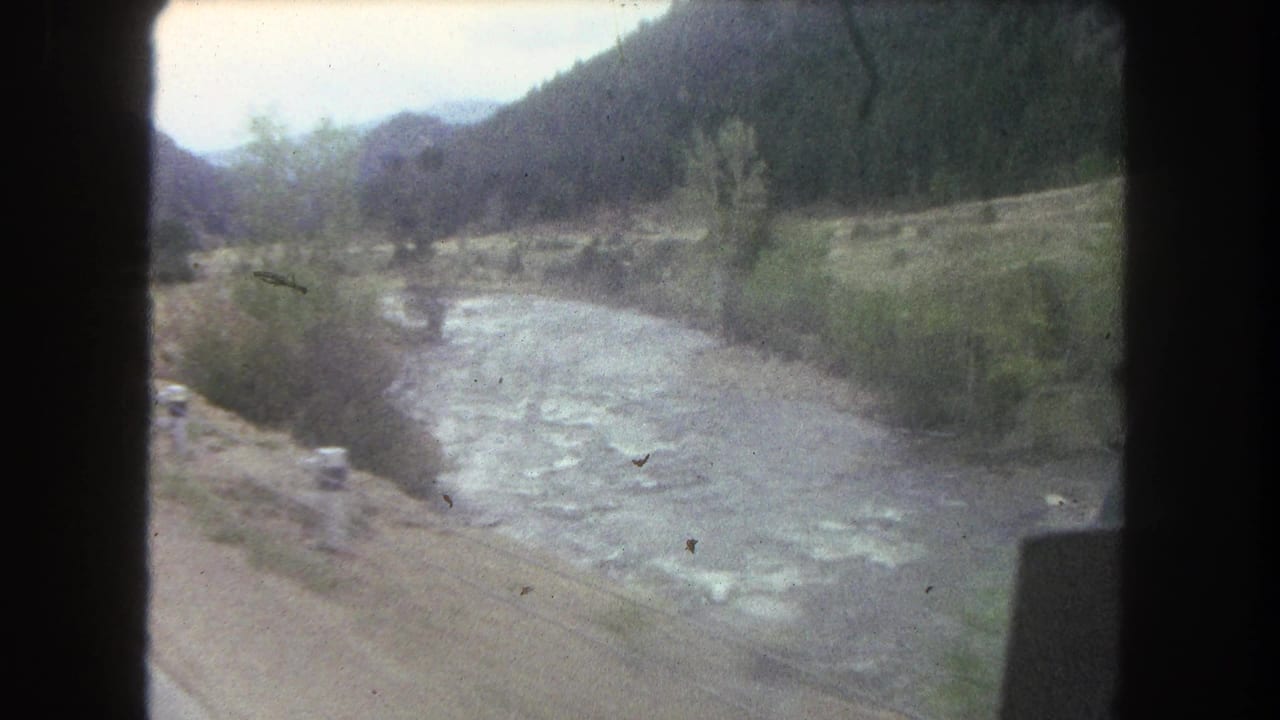 1962 Summit County, Colorado home-movie frame showing a river valley from a moving vehicle, with dirt road edge, trees, hills, and mountains.