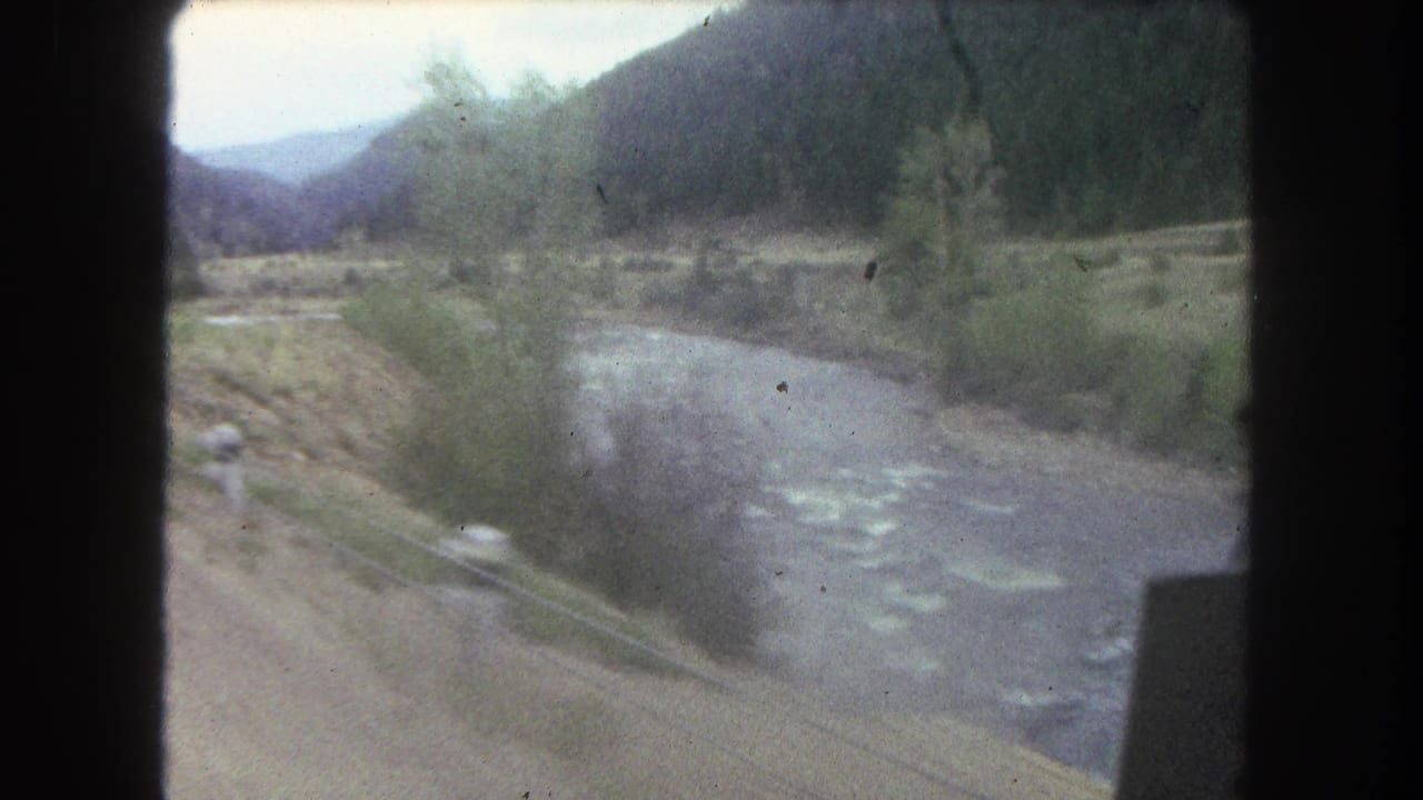 Moving-vehicle view in Summit County, Colorado, with a pale river, brushy banks, dirt shoulder, wooded slopes, and distant hills.