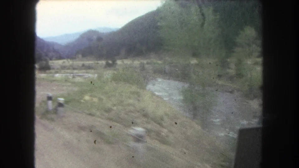 Soft 8mm frame from a 1962 Colorado valley drive, showing river water, meadow ground, roadside blur, leafless trees, and dark window edges.