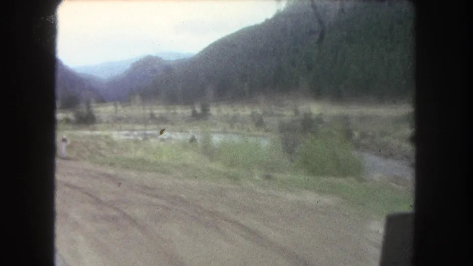 Summit County, Colorado home-movie view of open valley ground, dirt road tracks, a narrow river, wooded hills, and blue-gray mountains beyond.