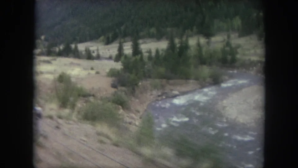 Passing 8mm frame from Winter Park, Colorado, with a river bend, brush, trees, and mountain slope viewed from a train.