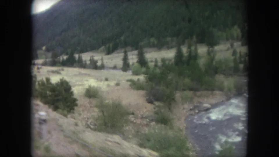 Soft 1962 8mm train-window view near Winter Park showing tracks, a river edge, shrubs, and forested Colorado hills.