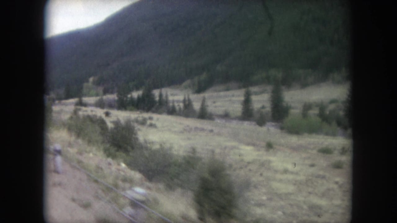 1962 Winter Park, Colorado frame with open meadow, roadside vegetation, evergreen trees, and a dark mountain slope beyond.