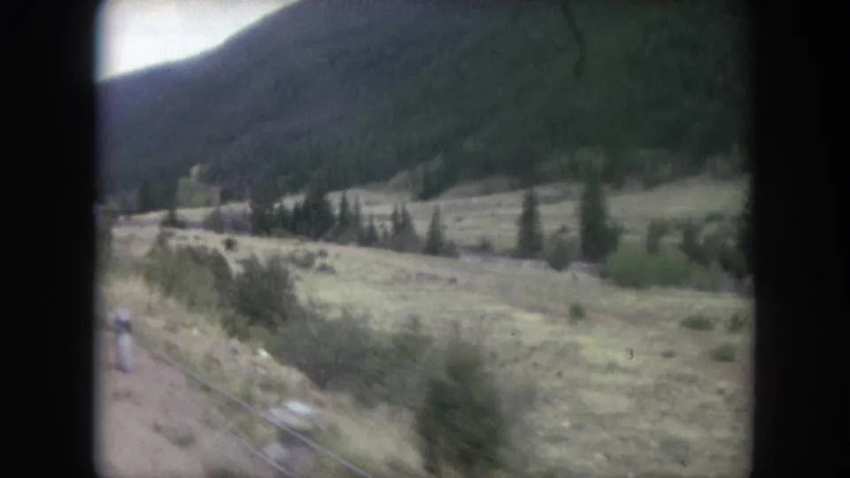 1962 Winter Park, Colorado frame with open meadow, roadside vegetation, evergreen trees, and a dark mountain slope beyond.