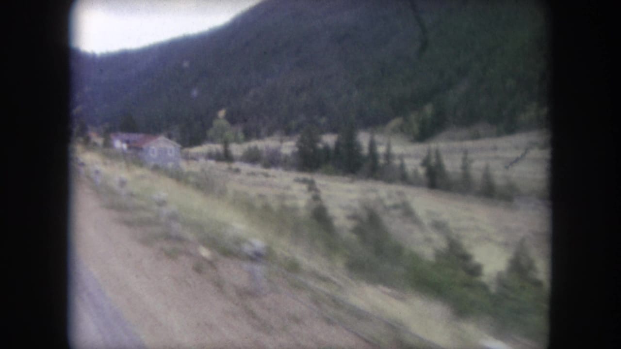 Vehicle-window view of a mountain road edge, grassy valley, pine forest, and distant hillside buildings.